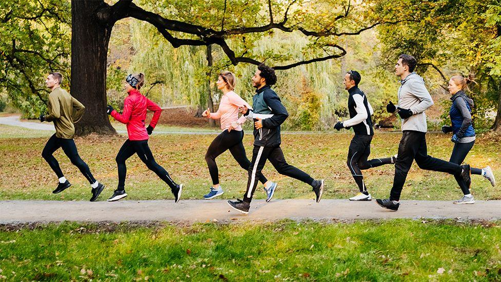 A group of seven joggers, of three females and four males, racing against each other on pedestrian walk way at the park, with green grass on either side and green trees in the background.