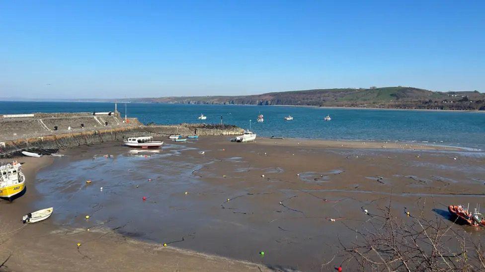 A view of Cardigan Bay, West Wales, UK. A curved sea wall is seen on the left, with boats in the harbour and water in the mid-distance.