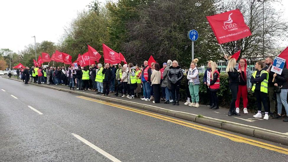 Striking workers holding flags stand on the pavement outside the Bakkavor factory