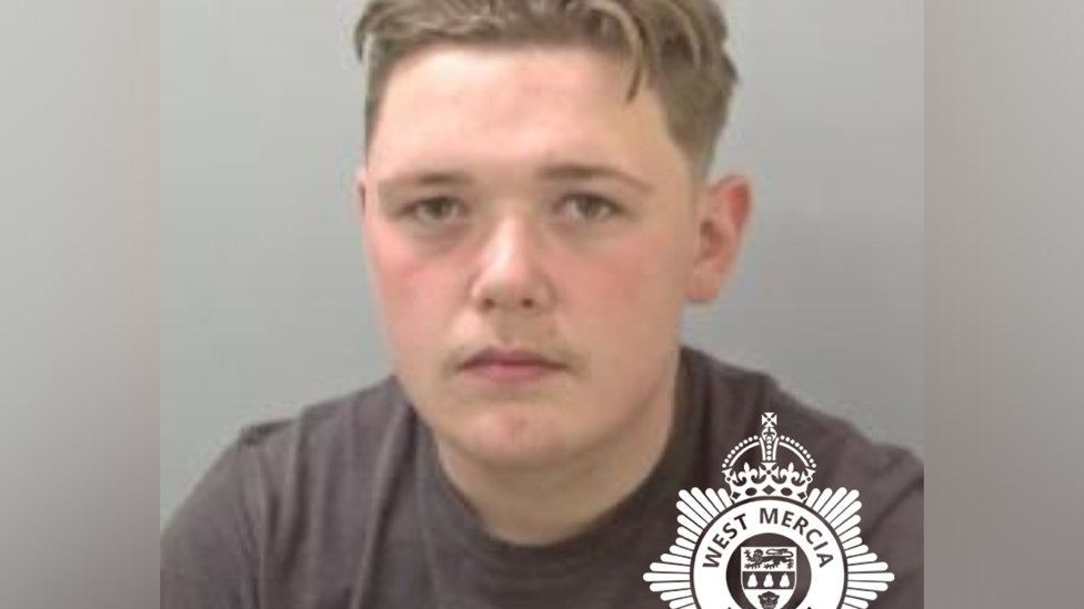 Mugshot of Logan Addison taken against a grey wall. He's wearing a black T-shirt and has a neutral expression.