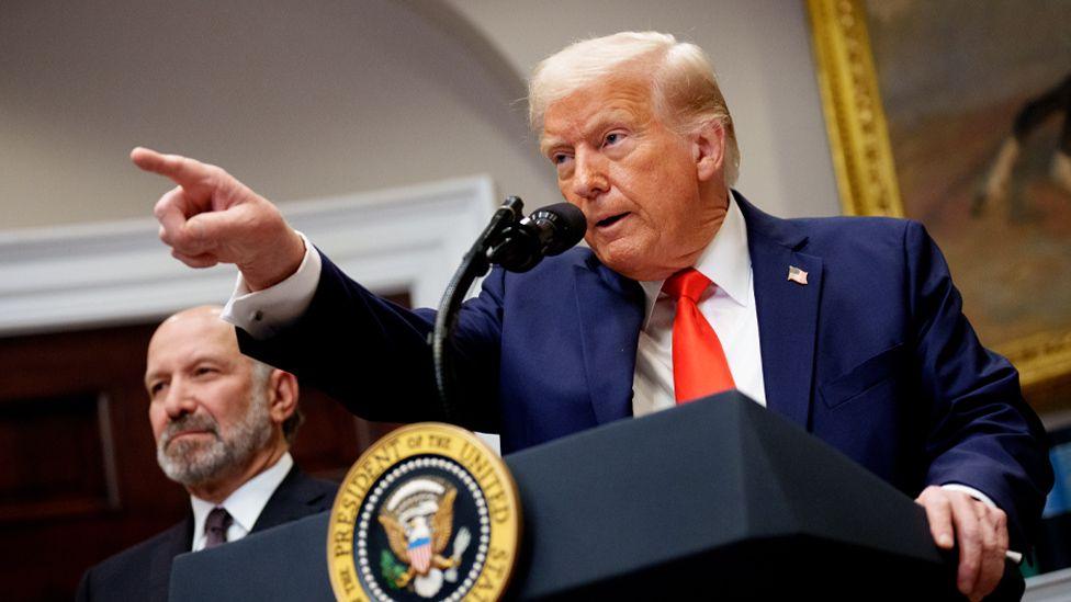 US President Donald Trump, wearing a blue suit and orange tie, pointing at an audience member while he speaks at a lectern 