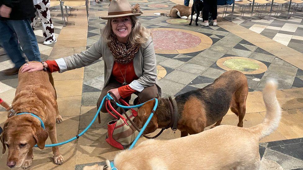 A woman wearing a grey jacket over a red jumper and a trilby-style hat, smiling at the camera and squatting down while holding three leads with three dogs attached in Ely Cathedral