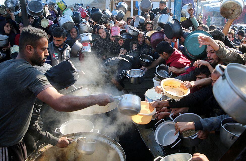 Palestinians jostle for space to receive food cooked by a charity kitchen. A man to the left ladles food into various receptacles held out by people in the crowd.