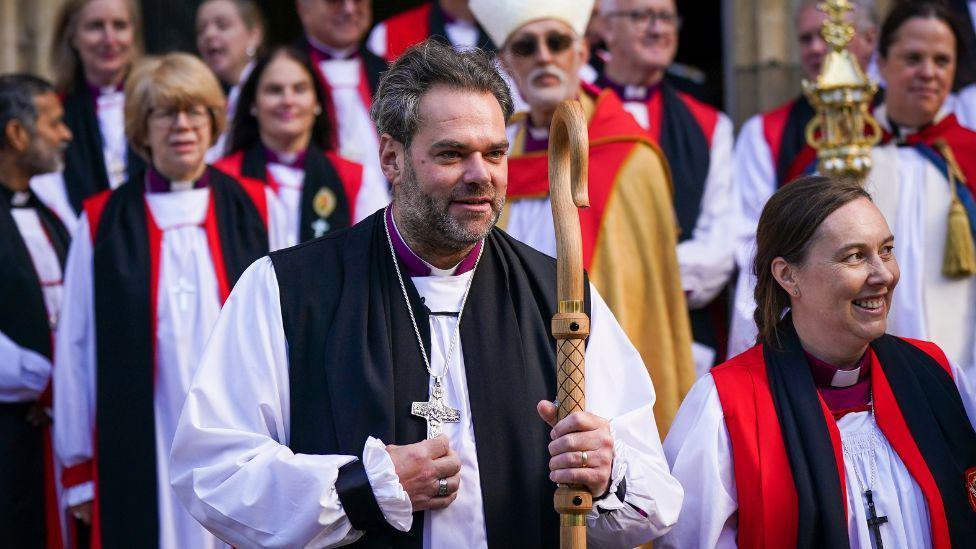 Barry Hill at York Minster. He is dressed in religious attire and is holding a large silver cross in his right hand.