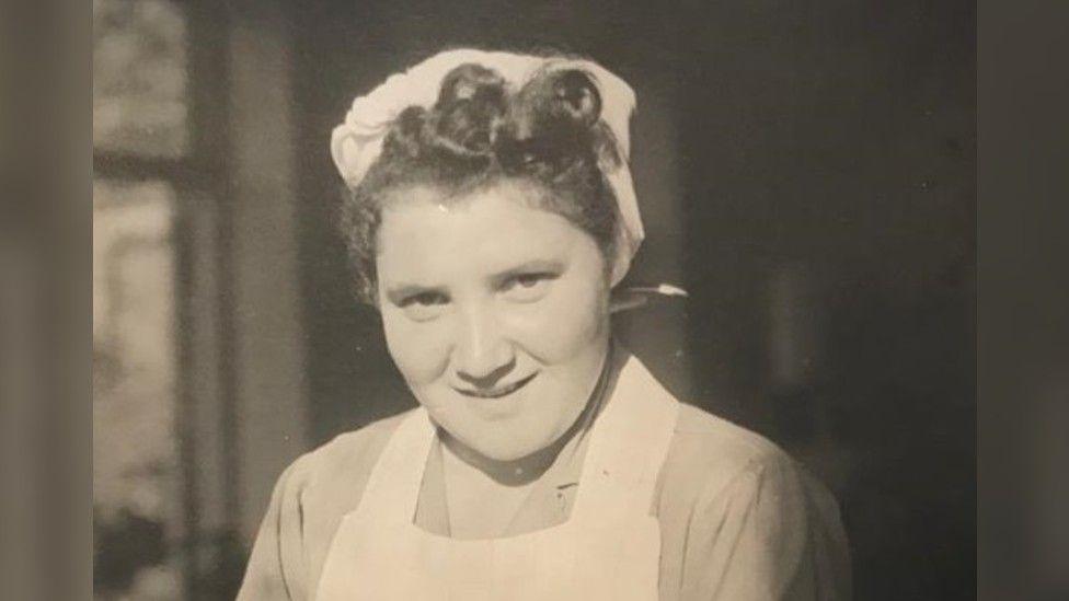 An old black and white picture of Mascha, taken as a young woman. She has pin-curled hair tucked under a white cap and is wearing a white apron over a light-coloured shirt. 