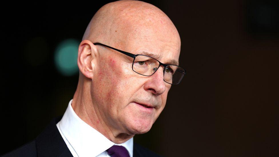 John Swinney, a bald man wearing glasses and a dark suit with a white shirt, speaks to the media in front of an out of focus black background 