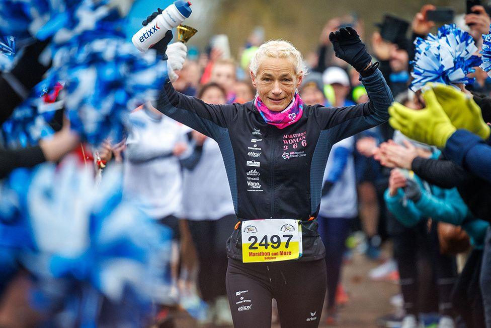 Marathon runner Hilde Dosogne, raises her arms as she finishes her 366th marathon of 2024 in Ghent, Belgium, on 31 December 2024.  She is wearing a pink neck-warmer and black and grey running outfit emblazoned with the  competitor number 2497. She has white/blonde hair and is framed by the  clapping hands of the crowd at the finishing line.