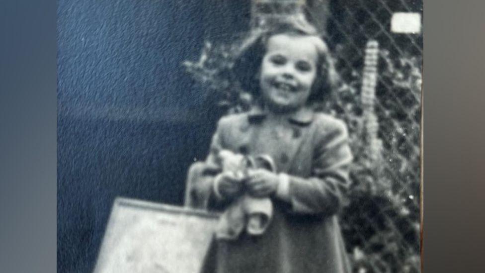 A black and white photo of Maggie Hackney as a young child. She stands outside in front of a wire fence, holding a pair of sandals and a shopping back. She has chin length hair and is smiling widely. She wears a smart duffel coat with a collar and double breasting.