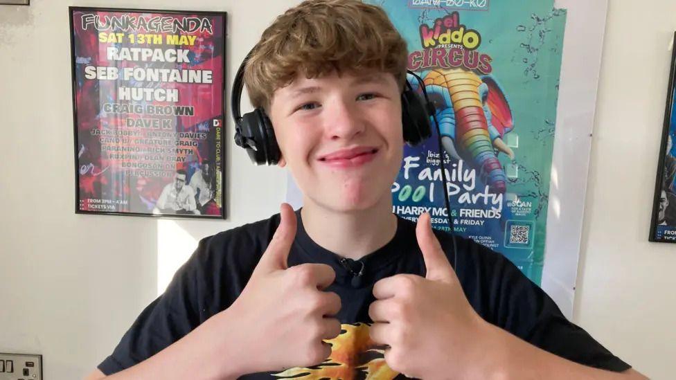 Jack Bobby standing in a room with posters on the wall. He is wearing a black t-shirt and headphones. He is smiling at the camera and giving the thumbs-up.