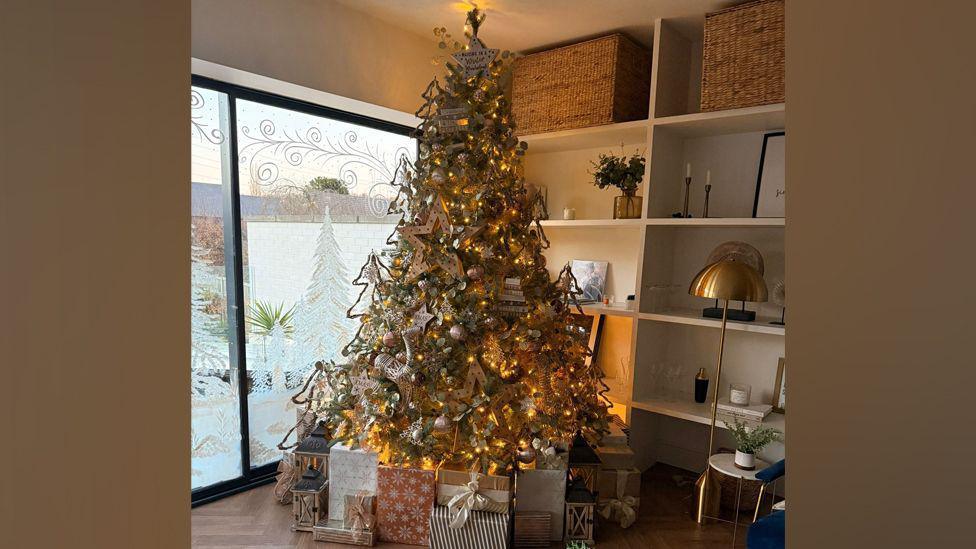 A Christmas tree lit up with large stars and lights. There are lots of beautifully wrapped presents under the tree. There is festive stencilling on some glass doors which lead to the garden behind the tree.