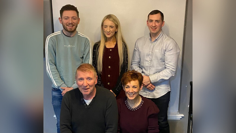 Family photo. Cahir is wearing jeans and a green jumper, Briege, with long blonde hair is wearing a burgundy dress, beside Paul in a grey shirt. In the foreground, sitting, is Kevin, with ginger hair wearing a dark top and mother Briege in burgundy top.