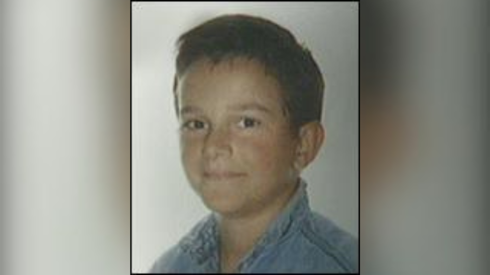 A young boy looking at the camera with a slight smile. He is wearing a blue denim shirt