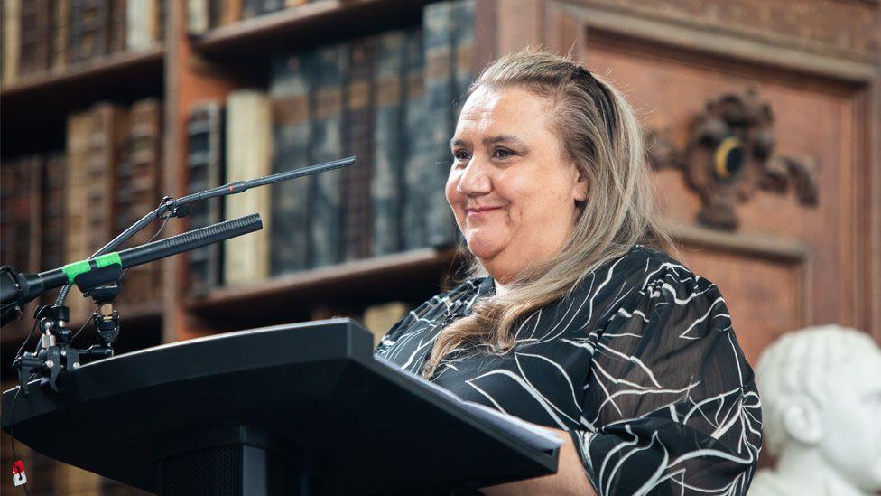 Noeleen Timbery standing at a lectern in front of a microphone in the Wren Library, Trinity College, Cambridge