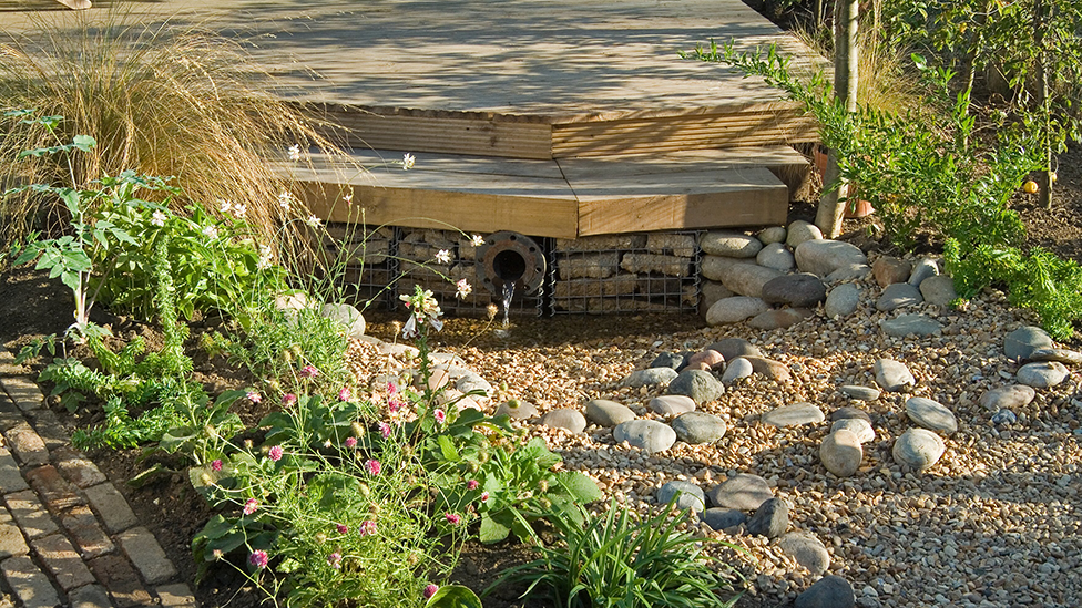 A garden with a gravel and stone section. Above it is a decking area, with planting to the left