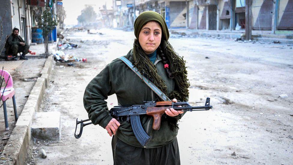 A woman holding a gun stands in a destroyed street in Kobane. There are wrecked buildings behind her.   