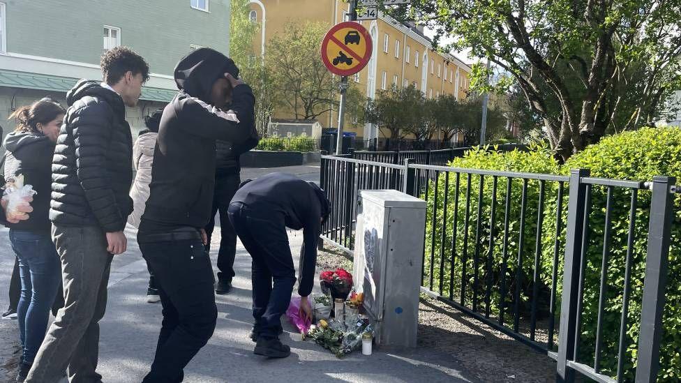 A group of people in dark clothing stand at a street corner. There is a pile of flowers on the ground and one person is laying more flowers down.