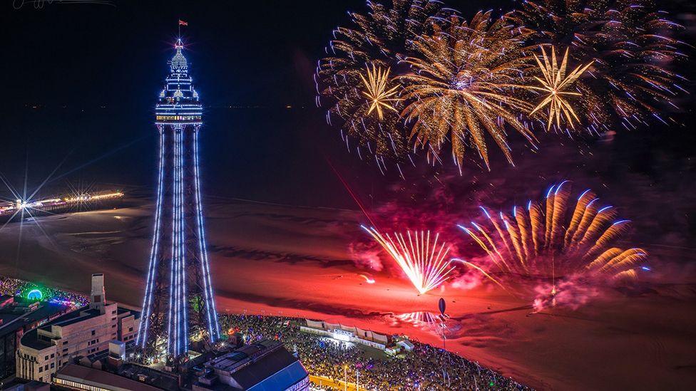 Aerial view of the fireworks display on the beach beside Blackpool's North pier with crowds of people watching on the promenade in the evening with the Blackpool Illuminations on and Blackpool Tower lit up purple in the background to the left.
