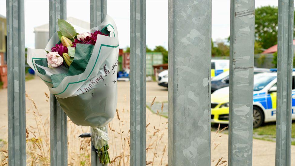 A close up of a bunch of flowers that has been strapped to the metal gate of the factory. It says Thinking of You on the bouquet and the name has been blurred out. Through the silver bars you can see a police car and police van parked alongside a black car.