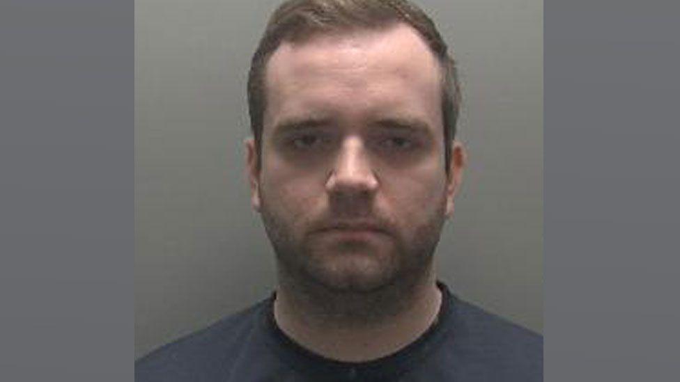 A slightly blurry police mug shot of Thomas Verdicchio, who has short light brown hair and stubble. He is wearing a dark blue T-shirt. 