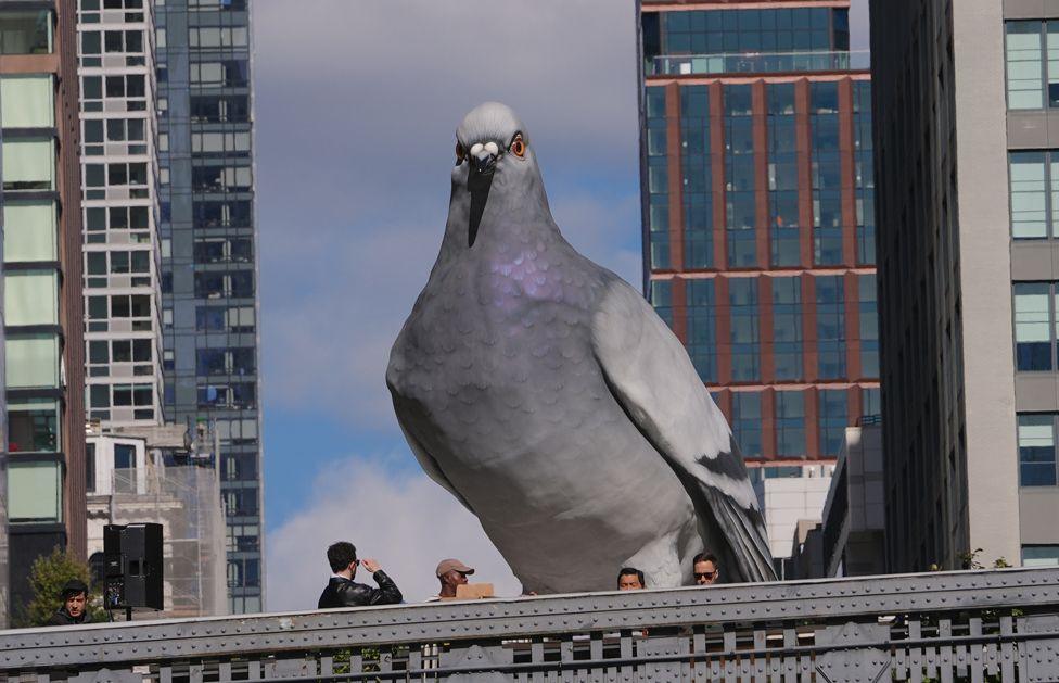 A large sculpture of a pigeon in the foreground with only the heads of people passing by visible below. Tall buildings are in the background and the sky is blue.