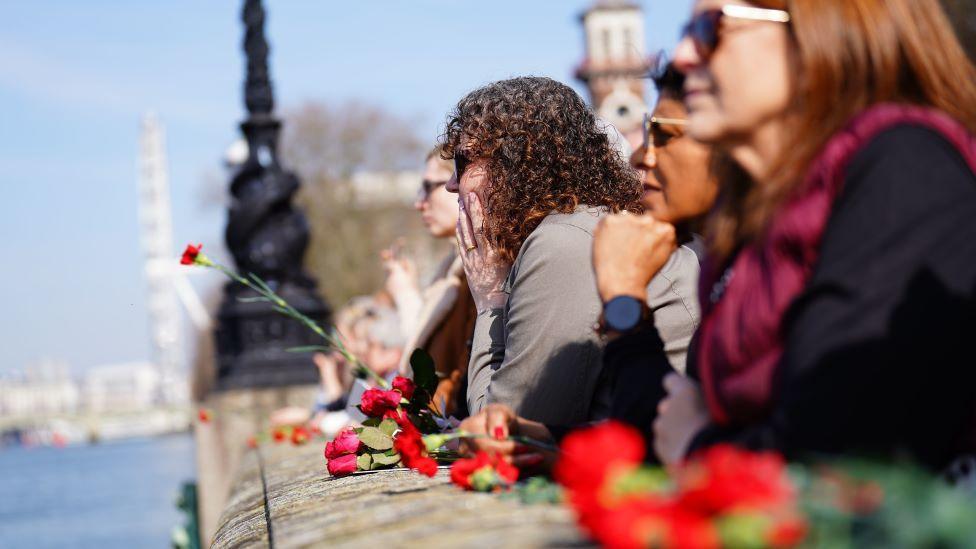 A row of women stand on a bridge that is strewn with red flowers