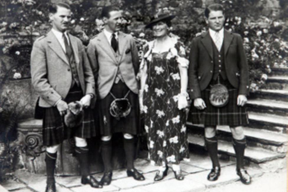 Black and white photo of three men in kilts, either side of their mother who is wearing a floral dress.