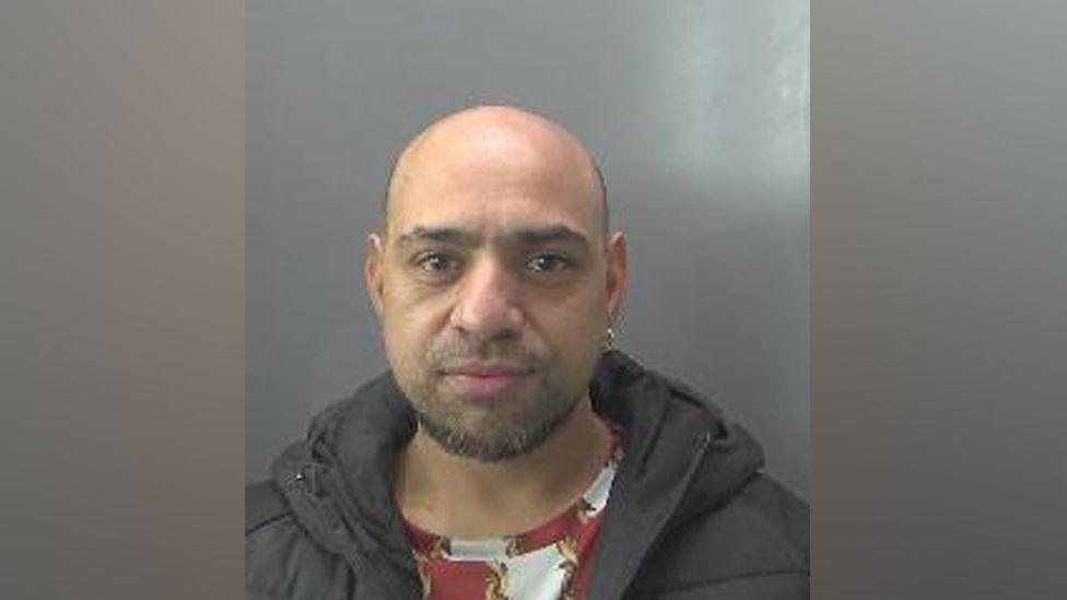 Mohammed Fiaz is looking at the camera in a police custody photo. He is mostly balding with a beard and moustache. He has an earring in his left ear. He is wearing a red patterned top and a black jacket
