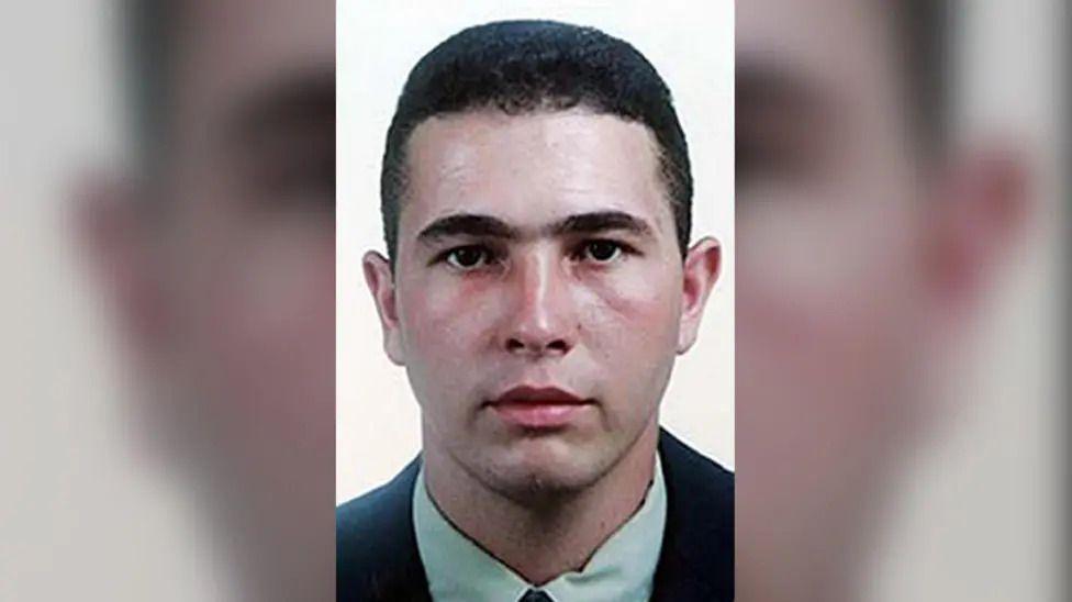 Passport-style photo of Jean Charles de Menezes. He has short dark hair and is wearing a green shirt and black jacket.