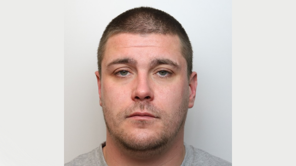 A mugshot of Grant Walker. He has short brown hair and grey-green eyes. He is wearing a grey t-shirt and standing against a grey background. He is looking at the camera with a blank expression.