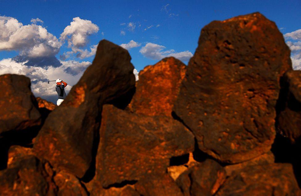 A male golfer is swinging a golf club in the distance, seen through slightly out of focus large dark rocks in the foreground under a bright blue sky with scattered clouds.