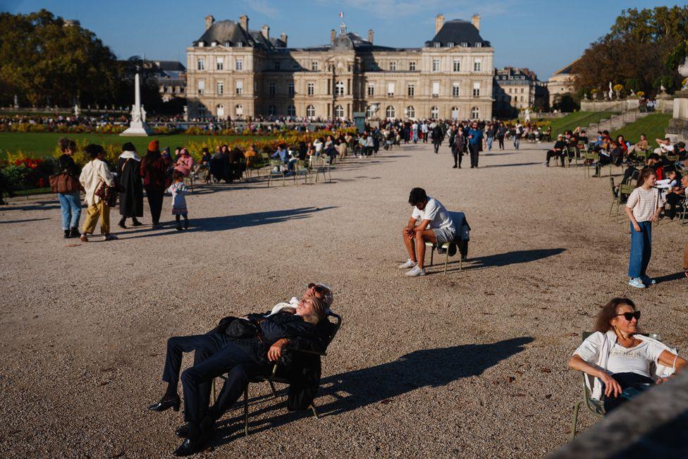 A woman lies on top of a man, both in black, lounging back on a chair in the foreground. One person in white sits on a chair behind them and a woman wearing white and sunglasses sits to the right. People walk along a promenade towards the Luxembourg Palace. Grass and trees line either site of the path.