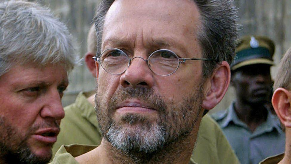 Simon Mann, the leader of the group of foreigners arrested in Zimbabwe for plotting a coup in Equatorial Guinea, is pictured in 2004 in a prison yard. He has wire-rimmed oval glasses on, sports a beard - and like the other prisoners seen in the background, is wearing khaki. An officer in a grey uniform with a peak hat can also been seen in the background.