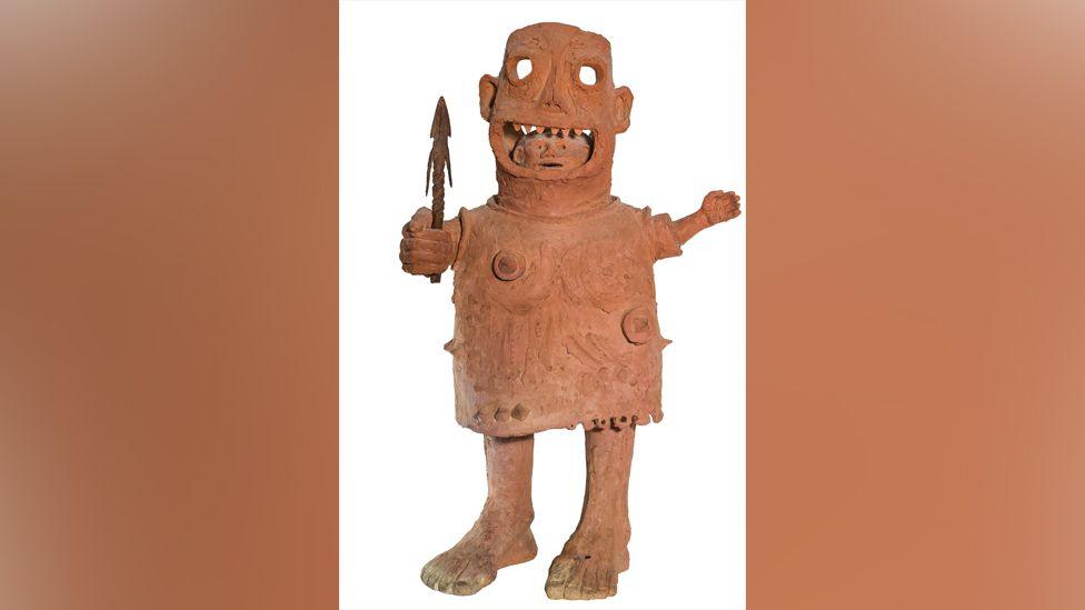 A clay sculpture shows a man's head staring out from a large mask. The creature depicted is wearing a tunic and holding a spear in one hand. The creature has very large feet