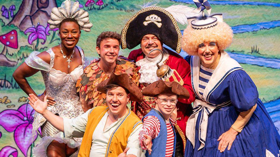 Top right Simone Robinson as Wendy Darling/Myrtle, in a silver dress and headdress; David Breeds as Peter Pan in a leafy costume; John Thomson as Captain Hook, with a large black hat, red jacket and holding up his left arm with its hook hand; Gordon Cooper as the dame in a curly blond wig, wearing bright make-up and a blue and white dress. Front row Oliver Scott as Smee, in a brown tricorne hat and yellow waistcoat over a white shirt; and Starky in a brown tricorne hat, red and white stripped top under a blue waistcoat, wearing clear plastic glasses.
