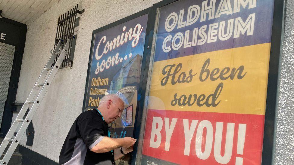 A workman installing a poster saying "Oldham Coliseum has been saved by you!"