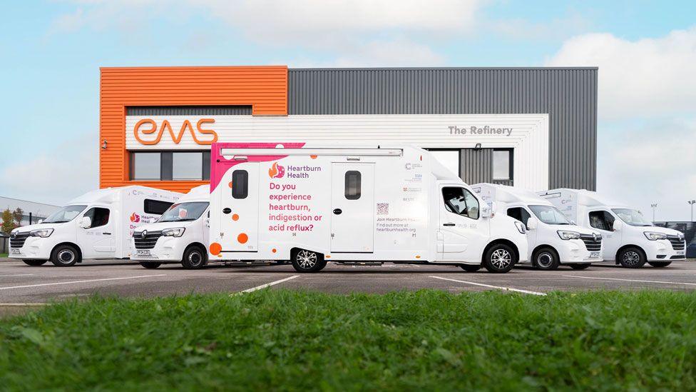 Five large white camper vans in front of a grey and orange building saying EMS. One of the vans is side on and it has pink writing saying heartburn health and do you experience heartburn, indigestion or acid reflux?