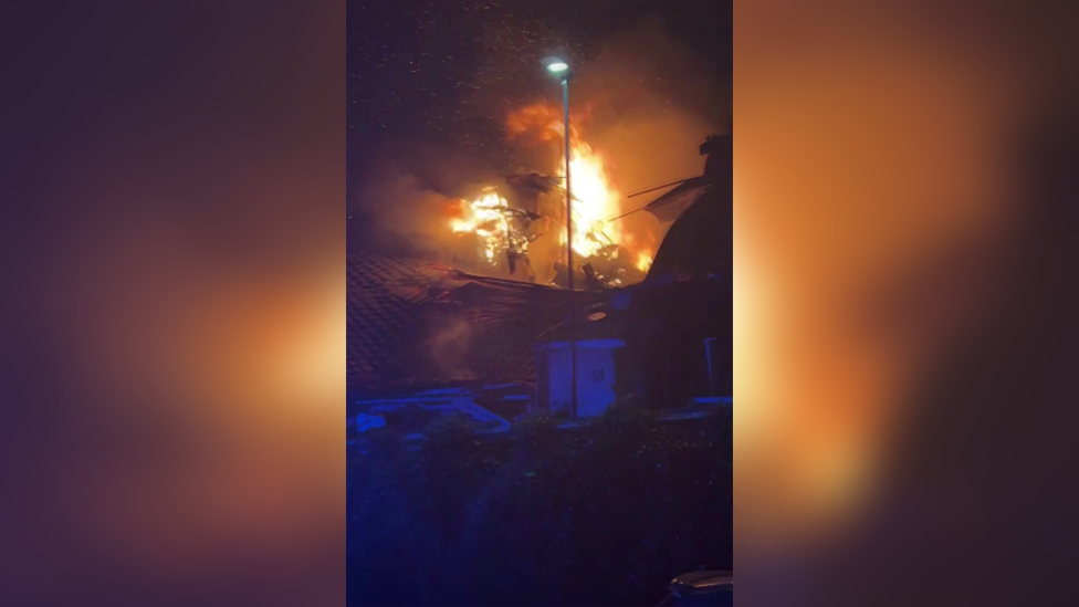 Picture shows large flames and a house that has been destroyed. It is nighttime.