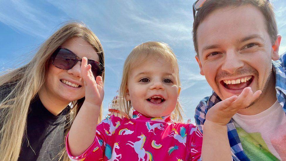 Rebecca Ableman (left), daughter Autumn (centre) and Chris Tuczemskyi (right) are all looking down at the camera on a sunny day with blue skies behind them. Rebecca has long blonde hair and sunglasses. Autumn is holding her hands up and is wearing a pink top with rainbows and unicorns on. She has blonde hair. Chris is also smiling at the camera and is wearing a blue checkered shirt with a white top. He has short brown hair and sunglasses on his head.