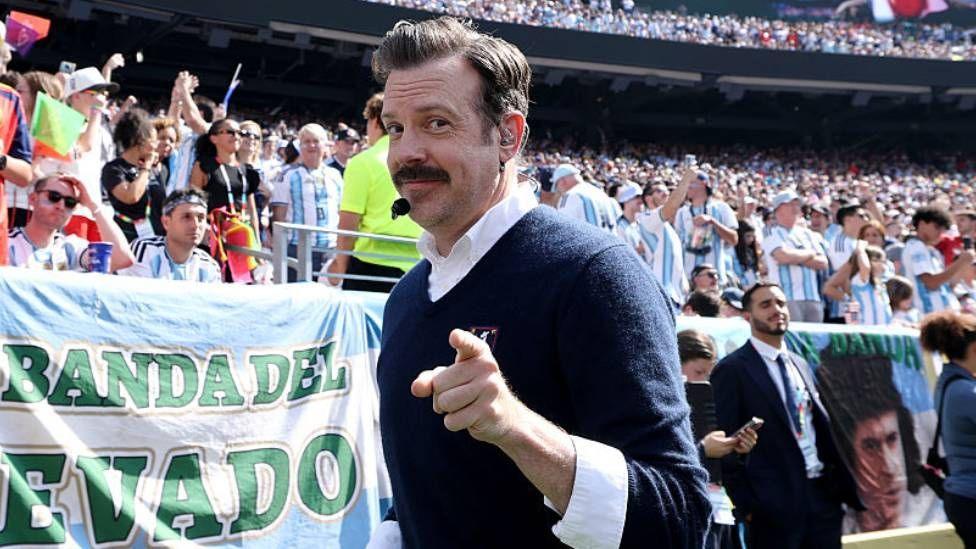 Ted Lasso seen at a football match, wearing a blue jumper over a white shirt