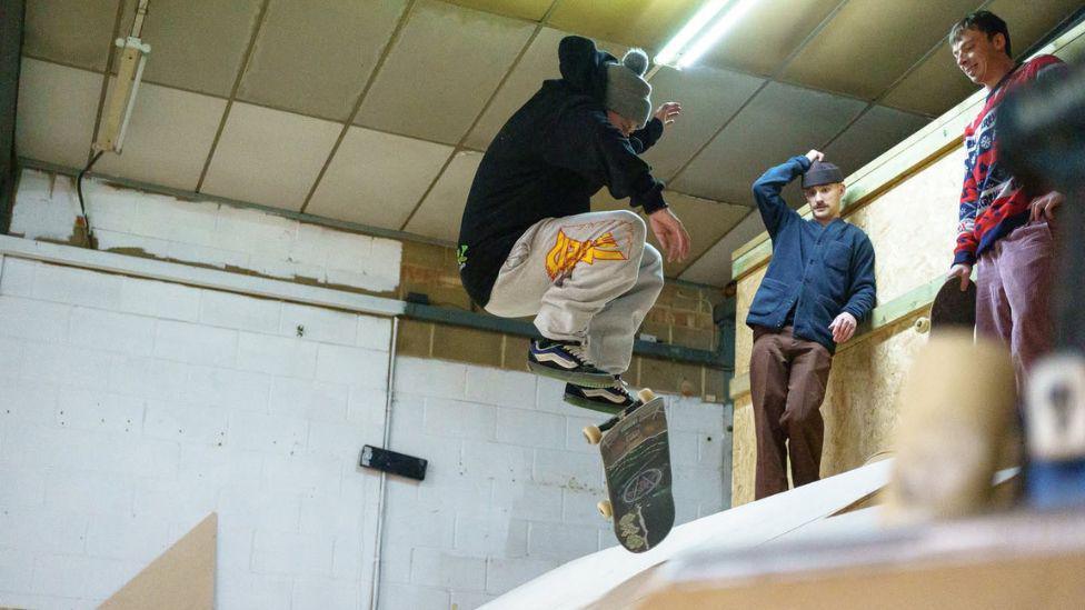 A man doing a trick on a skateboard down a ramp. He is wearing a woolly hat, a black jumper and grey trousers. There are two more men stood at the top of the ramp behind him.