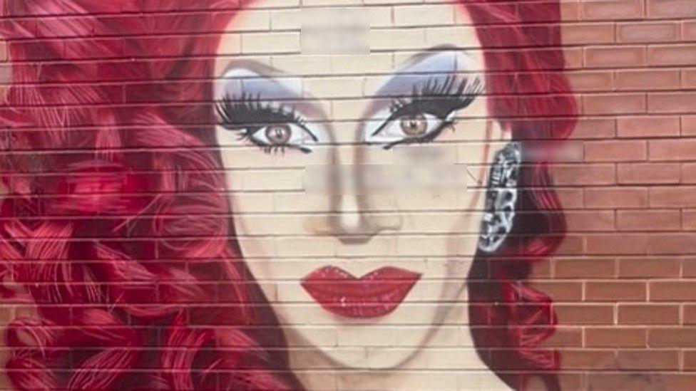 A colourful mural of drag queen Divina De Campo, with red curly hair and red lipstick, with sections where the mural has been defaced by graffiti blurred out