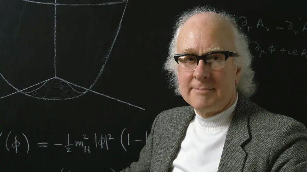 Prof Higgs is in front of a black board with calculations on it. He has white shoulder-length hair and black rimmed glasses. He is wearing a tweed suit jacket with a white polo-neck jumper underneath.