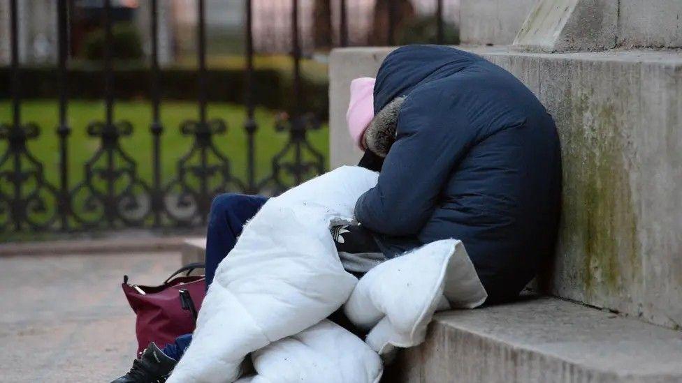 A person sitting on a concrete bench wearing a blue hooded jacket with a white quilt wrapped around them.