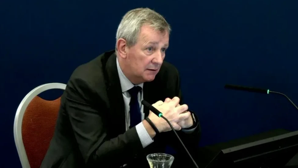 Richard Pengelly. He has short grey hair, wearing a black blazer, white shirt and black tie. He is sitting in front of a microphone and his hands are clasped together.