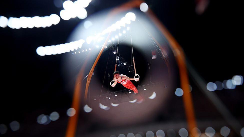 Gymnast in a red uniform performs on the rings under bright arena lights, suspended mid-air with blurred lights creating a dramatic effect in the background.