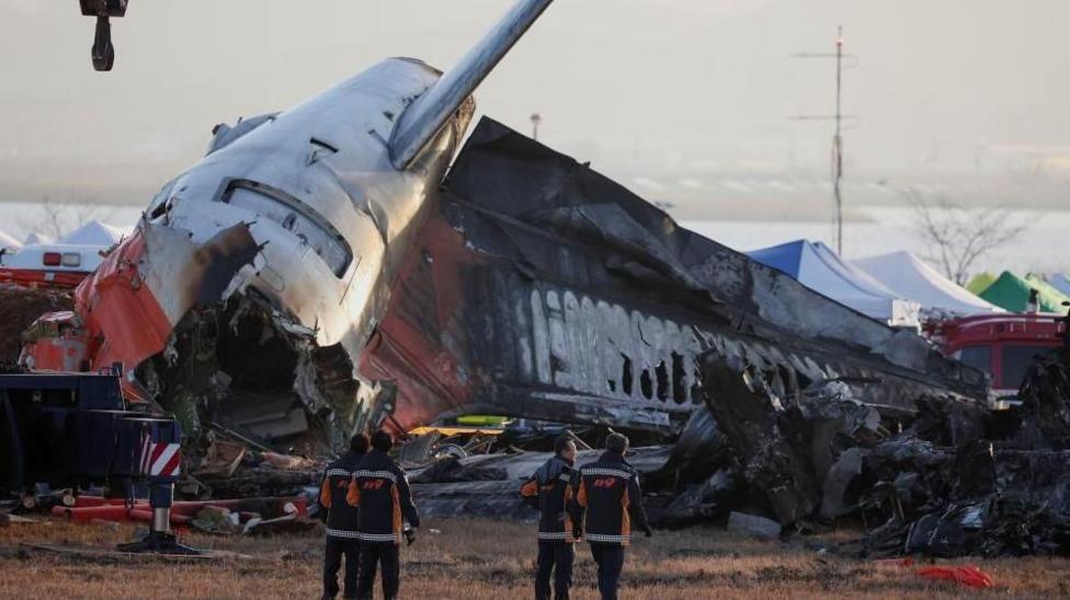 Wreckage of the crashed Jeju Air jet. Four firefighters wearing dark clothing are looking up at it with their backs to the camera