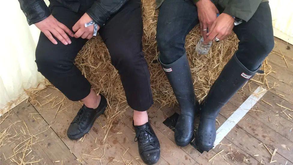 Footwear of Feeder band members Taka Hirose, left, and Grant Nicholas. Taka is wearing black shoes and Grant a pair of wellies. The musicians are sitting on a bale of straw and there is straw on the wooden-board floor below.