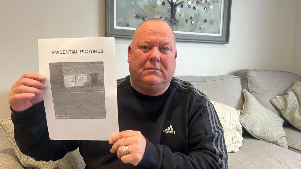 Damian Quillan sat on a sofa holding a piece of paper which says "evidential pictures" on it