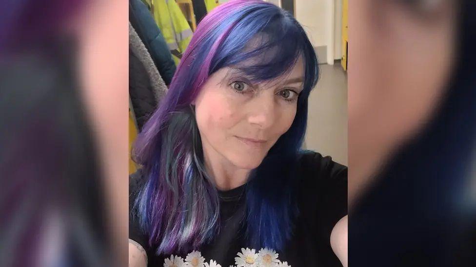 Rebecca Ashmead pictured in a selfie. She has distinctive blue and purple hair, cut into a fringe, and wears a black t-shirt with white daisies on it. 