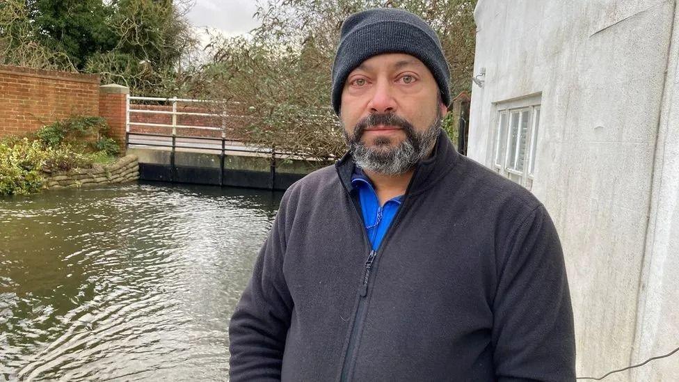 A bearded man wearing a black fleece and a beanie hat stands in front of a river, the level of which has risen to the underside of a bridge